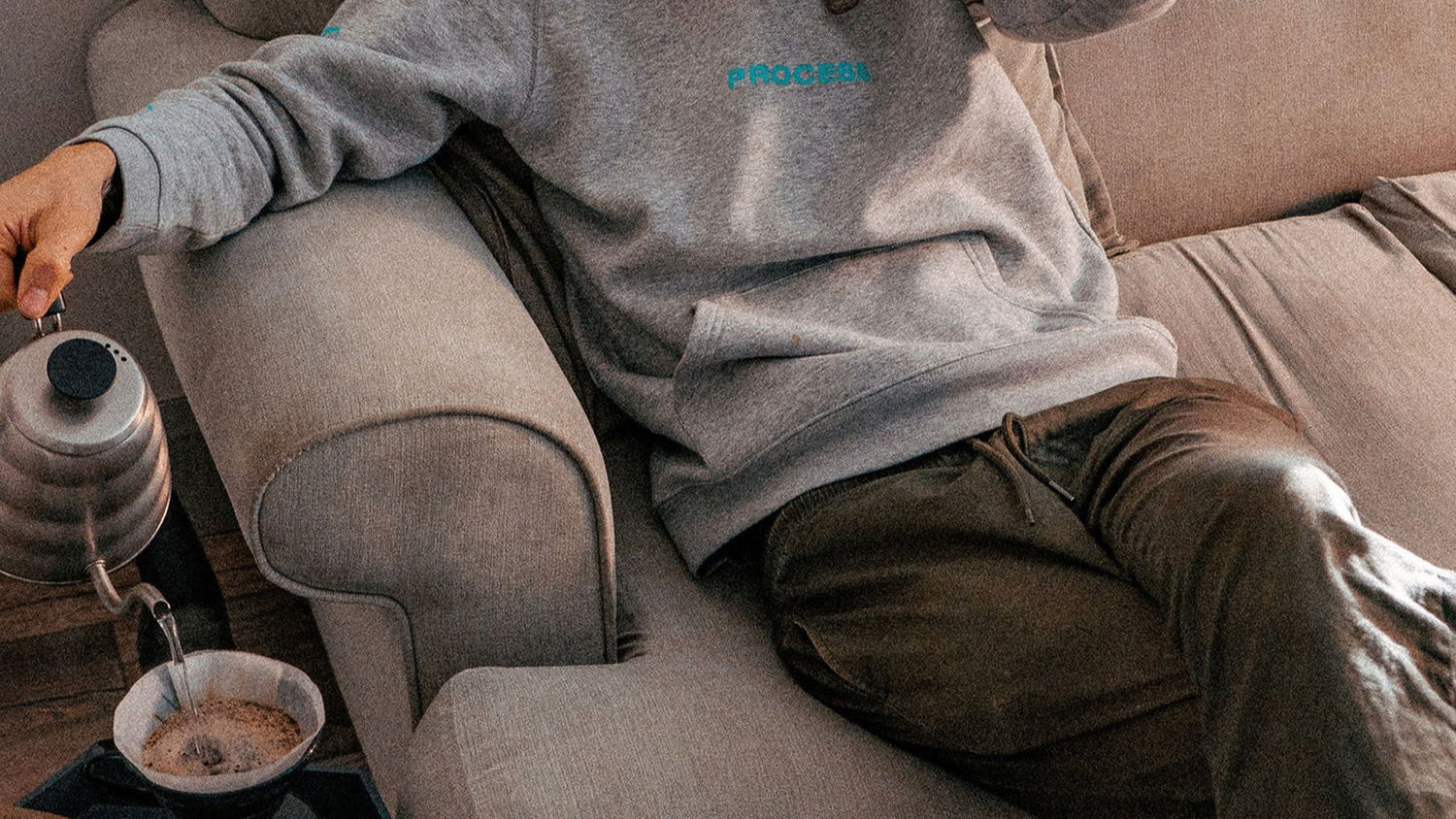 Person sitting on a couch pouring coffee and wearing a grey sweatshirt with Process Coffee logo.